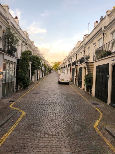 A narrow cobblestone street in Marylebone with white terraced houses on both sides, each featuring black or dark-colored garage doors and small front gardens decorated with greenery and potted plants. A white van is parked on the right side of the street, close to the curb, with its rear doors partially visible, indicating it is in the process of being loaded or unloaded. The street is lined with yellow double parking restriction lines, and some houses have balconies with iron railings. In the distance, the street gently slopes upward, illuminated by soft natural light as the sky above shows a mix of clouds and clear blue. The scene captures a typical residential area ideal for house removals or furniture transport involving careful loading and unloading within a tight urban environment, a process often managed by services like Man and Van Marylebone.