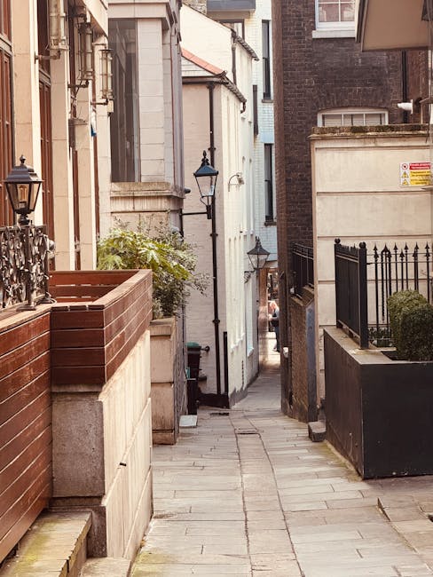 A narrow residential alleyway in Marylebone during daytime, with paving stones and tall brick buildings on either side. On the left, a wooden planter box is positioned atop a concrete ledge, accompanied by black iron railings and small outdoor lamp posts attached to the building. On the right, a black metal fence surrounds a small garden area with a neatly trimmed shrub, and a yellow warning sign is attached to the upper part of the building's wall. The alley's view extends towards the background, where a vehicle is parked at the end, and the scene suggests preparations for home relocation or furniture transport involving loading and unloading processes, supported by the proximity to nearby properties and typical urban street environment, indicating professional removals potentially carried out by companies like Man and Van Marylebone.