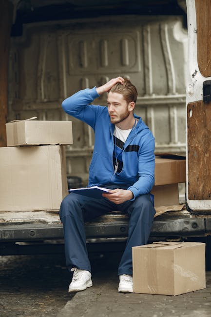 A man wearing a beige knit beanie, a green and navy puffer jacket, and black gloves is sitting on the pavement in front of an open van during a home relocation. Inside the van, several cardboard movin