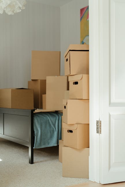 A collection of cardboard moving boxes of various sizes stacked on top of each other and placed on a bedside table inside a room, near a partially open white door. The boxes are sealed with packing tape and some have cut-out handles. The room has light-colored walls and a colourful painting hanging on the wall behind the storage. Natural light illuminates the scene, highlighting the boxes prepared for a home relocation. The stacked boxes are ready for transport, with some secured with plastic wraps, and are situated close to a bed draped with a light blue bedspread. This setup illustrates the packing and loading process typical of house removals services provided by Man and Van Marylebone, focusing on handling bulky items and ensuring safe furniture transport without damage.