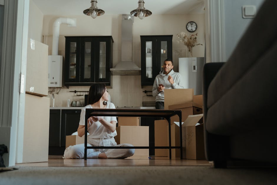 Inside a residential kitchen area with cream-colored walls and ceiling-mounted pendant lights, a woman with dark hair is sitting cross-legged on the wooden floor, holding a smartphone and smiling. Surrounding her are several packed cardboard boxes of various sizes, some sealed with packing tape, indicating a home relocation process. Behind her, a man wearing a light grey hoodie is standing near the counter, holding a mobile device, possibly coordinating the moving process. The kitchen features black wall-mounted cabinets with glass doors, a stainless steel oven hood, and a wall clock indicating the time. To the right, part of a grey sofa is visible, and on the left side, there are white appliances, including a microwave and what appears to be a water heater. The scene captures the preparation and packing stages of furniture transport, with the interactions and environment reflecting a professional moving service like Man and Van Marylebone handling logistics for household relocation and packing.