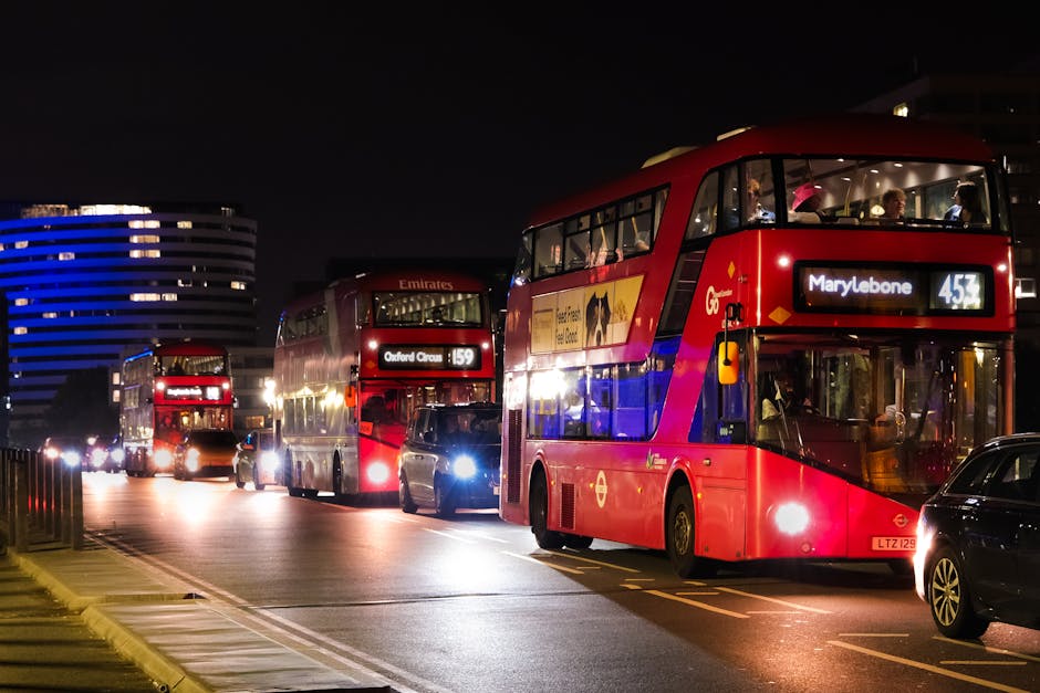 Nighttime scene featuring multiple red double-decker buses parked along a city street near Marylebone, with visible front and side windows illuminated from inside. The buses display route information, including one marked 'Marylebone' and another 'Oxford Circus,' indicating ongoing bus routes relevant to residents or visitors. The street is lined with parked cars, and a few streetlights cast a subtle glow on the pavement. In the background, modern office buildings are lit with blue lights, creating a contrast against the dark sky. The scene captures the urban environment typical of a busy London route, possibly near Marylebone station, demonstrating transport options that could be included in house removals, furniture transport, or home relocation logistics managed by [COMPANY_NAME]. The image reflects the urban setting for moving or storage services, emphasizing loading or parking areas close to residential or commercial premises, with a focus on the nighttime traffic and bus transit infrastructure.