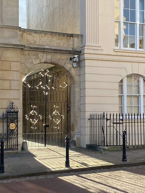 Photograph of the exterior entrance to a historic building during daylight hours, featuring an ornate black wrought iron gate with decorative scrollwork, positioned within a stone archway. The building’s facade is constructed of beige stone with classical architectural details, including tall, fluted pilasters, large sash windows, and a smooth finish. The sunlight casts shadows on the pavement and creates reflections on the gate's metal surfaces. A security camera is mounted above the archway, indicating monitored access. Two short black metal bollards are installed on the pavement in front of the gate, providing a barrier for pedestrian safety. The scene captures the quiet street environment in Marylebone, with the gate potentially serving as an entrance to a private residence or reception area. This setting is relevant for house removals and home relocation services provided by Man and Van Marylebone, illustrating a typical urban entryway involved in furniture transport and moving logistics.