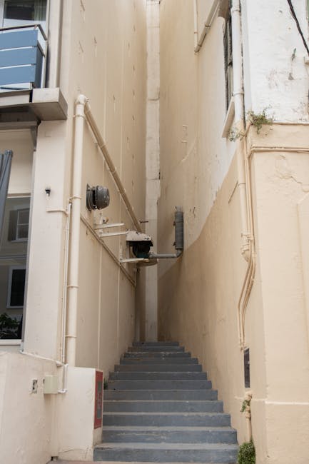 A narrow outdoor staircase with dark grey steps is situated between two beige building walls, leading upwards out of view. The left wall features visible white PVC pipes, an electrical box, and a black conduit, while the right wall has some minor plant growth and a vertical pipe. The area is lit by natural daylight, casting soft shadows. This outdoor environment is part of a property in Marylebone, and the scene relates to the home relocation process, where careful luggage or furniture transport through limited access points might be necessary. The image supports the context of London house removals and moving logistics handled by Man and Van Marylebone, illustrating challenges faced during property access and stair navigation in tight urban spaces used in furniture transport and packing and moving activities.