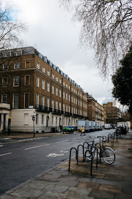 A multi-storey residential building with a brown brick exterior and white window frames, situated on Marylebone High Street, with the street scene including parked bicycles secured to black metal bike racks on the sidewalk. A cyclist in yellow attire bikes past, and several delivery trucks are parked along the curb, with one truck's rear doors open, indicating possible loading or unloading activity related to home relocation or furniture transport. Outside, there are some pedestrians, and the overcast sky casts diffuse light over the scene. The environment suggests an urban setting suitable for professional removals services, such as those provided by Man and Van Marylebone, which supports packing, moving, and furniture transport during house relocations, with visible materials like cardboard boxes and protective blankets possibly used for moving household items inside the property or during loading into the vehicles.
