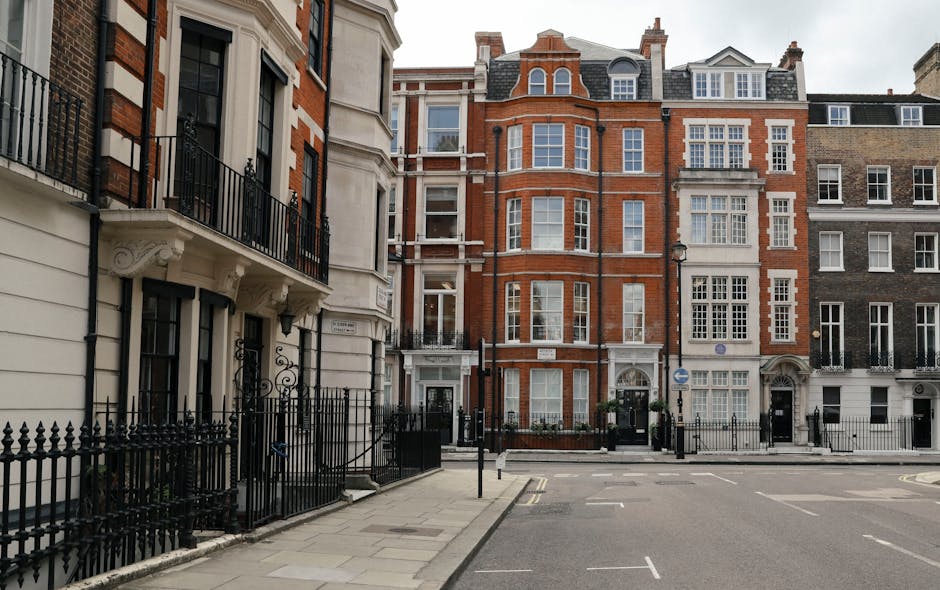 Street view showing a row of multi-storey residential buildings in Marylebone, constructed with brick and stone facades, with large sash windows and decorative architectural details. The scene includes a sidewalk with black metal fencing along the front of the buildings, and a narrow road with parked cars and a one-way traffic sign. The area appears quiet and well-maintained, suitable for a house removal or home relocation in Marylebone. This image exemplifies the urban environment where Man and Van Marylebone conducts furniture transport and packing and moving services, supporting efficient moving logistics in a residential setting.