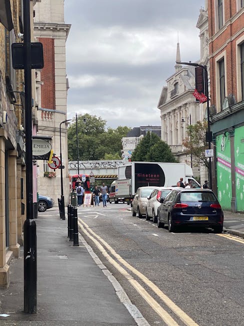 A narrow street in Marylebone, London, with parked cars along the curbside, including a black hatchback close to the foreground. The street is lined with historic red-brick and white stone buildings on both sides, some with shopfronts and signage. In the distance, a white delivery van from Man and Van Marylebone is being loaded or unloaded, with people nearby handling large cardboard boxes and furniture wrapped in protective plastic or blankets, indicating active furniture transport as part of a home relocation. The scene is under an overcast sky, and a double yellow line runs along the edge of the road, typical of UK parking regulations. The overall setting suggests a professional moving or packing operation in an urban residential area, with the service process underway for a same-day removals job.