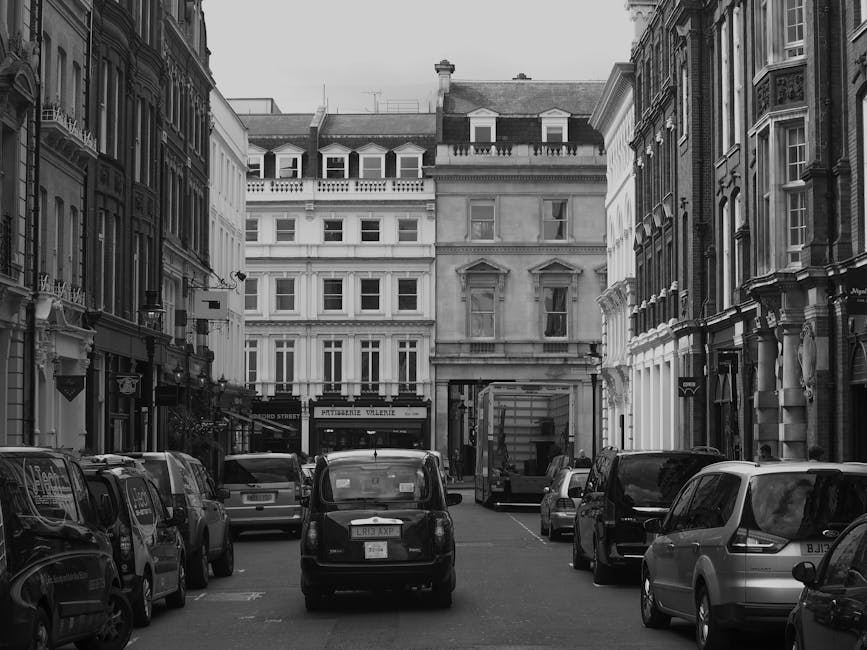 Black and white photograph of a narrow urban street in Marylebone, lined with multi-storey Victorian-style buildings with decorative facades and large sash windows. Several parked cars, including a small black cab and various hatchbacks and vans, occupy the street's curbside parking areas. On the right side, a delivery truck is seen partially extending into the street, indicating a loading or unloading process. In the background, the street opens to a small square or junction, with a mix of residential and commercial properties, including a visible signage for a restaurant or cafe. The scene suggests a typical city environment suitable for home relocation and furniture transport services, with the vehicles and surroundings reflecting logistics involved in packing and moving operations, which might be coordinated by companies like Man and Van Marylebone.