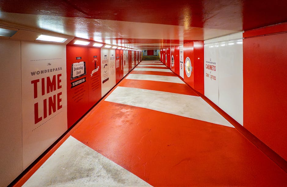 A long underground corridor with a predominantly red and white colour scheme, featuring a shiny red ceiling and red lower walls, with white upper walls decorated with various posters and signs, including one that reads 'The Wonderpass TIME LINE'. The floor has a pattern of alternating red and white diagonal sections, creating a dynamic visual effect. The corridor is lined with lockers or storage cabinets on the left side, and it extends into the distance, illuminated by evenly spaced rectangular ceiling lights. This setting suggests a storage or loading area within a building, with space for moving and handling containers or boxes, relevant to house removals and furniture transport services offered by Man and Van Marylebone.