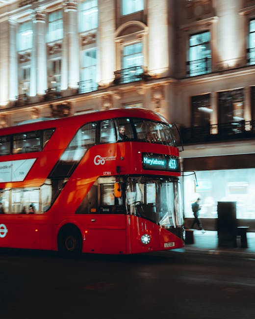 A street scene in central Marylebone featuring a bright red double-decker London bus with the destination display reading 'Marylebone.' The bus is parked or moving along a roadway in front of a historic building with ornate architectural details, large windows, and decorative columns. Pedestrians are visible walking on the sidewalk near the bus, and the bright natural lighting suggests daytime. The surroundings imply an urban environment typical of London, relevant to house removals and furniture transport services offered by Man and Van Marylebone as part of home relocation and moving logistics.