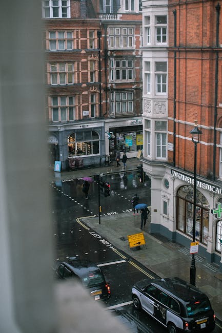 A view of a busy street in Marylebone during rainy weather, seen from a high vantage point through a window frame. On the street, several pedestrians with umbrellas walk along the pavement, while cars, including a black van and a black sedan, are parked along the curb. The scene features historical red brick buildings with bay windows and white framing, typical of Marylebone architecture. A small shop with signage and a pharmacy with a green cross are visible on the ground floors. The wet pavement reflects the streetlights and storefronts, and a yellow road sign indicates parking restrictions. This setting illustrates typical urban activity related to home relocation and furniture transport, with external conditions highlighting the need for careful planning during moving logistics, which [COMPANY_NAME] may assist with for compliance with Westminster Council permits for removals in Marylebone.