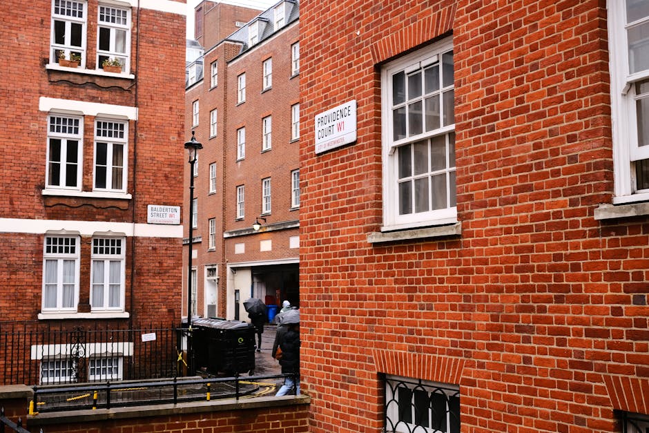 View of a narrow urban street corner in Marylebone, London, with red brick residential buildings featuring white framed sash windows. A street sign for 'Providence Court W1' is mounted on one building, and another for 'Balderton Street W1' is visible at the intersection. The scene shows part of a home relocation process by Man and Van Marylebone, with a black van parked on the street, partially visible, and two movers dressed in grey jackets carrying large cardboard boxes and a piece of furniture towards the van. The street area includes a small fenced section with a black metal gate, a waste disposal bin, and a portable yellow barrier. Overcast lighting casts soft shadows, emphasizing the typical London residential environment, and the surroundings suggest preparation for furniture transport or packing during a house removal.